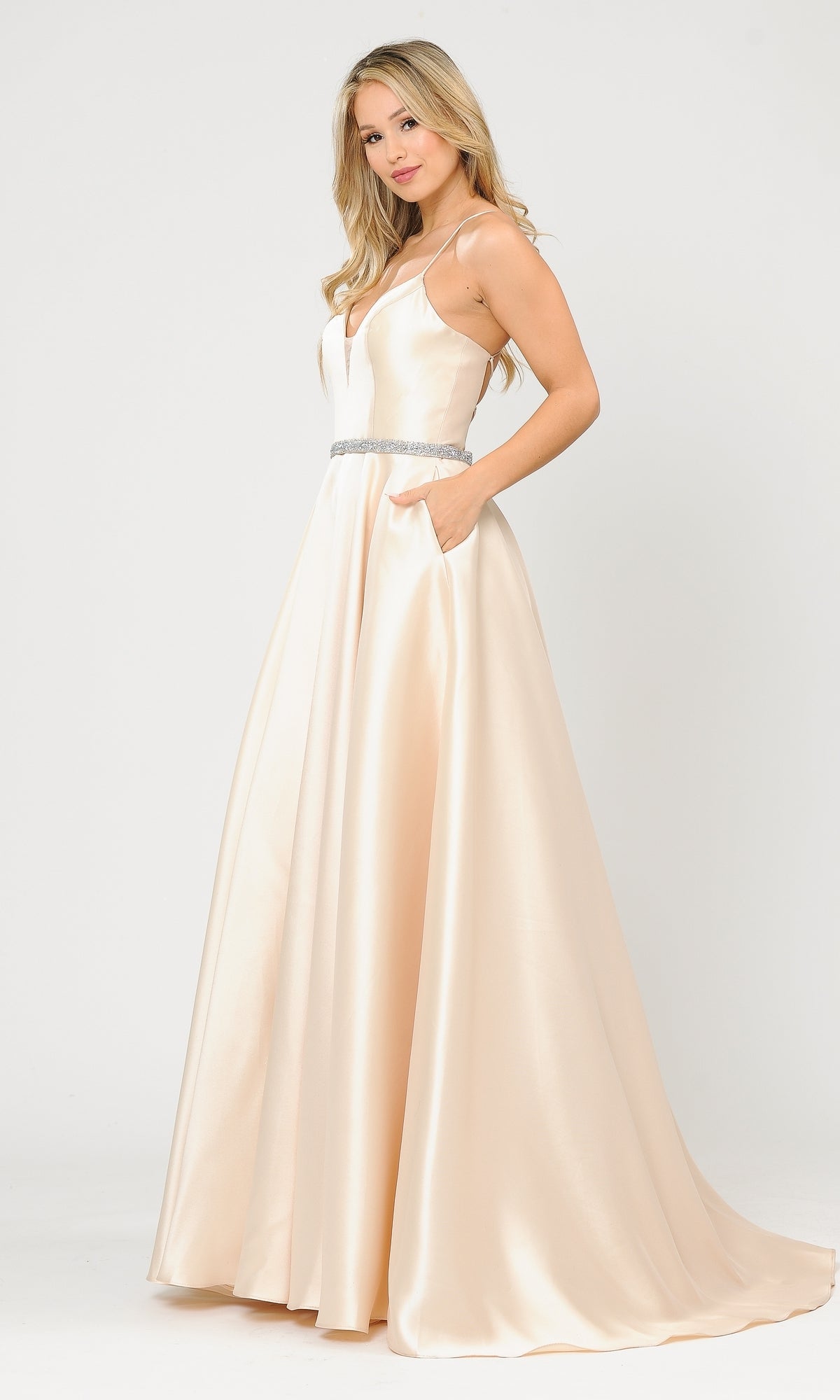 A woman wearing the Strappy-Back Long Mikado A-Line Prom Dress 8688—a sleeveless champagne gown with a V-neck, fitted bodice, jeweled belt, and side pockets—stands against a plain white background.